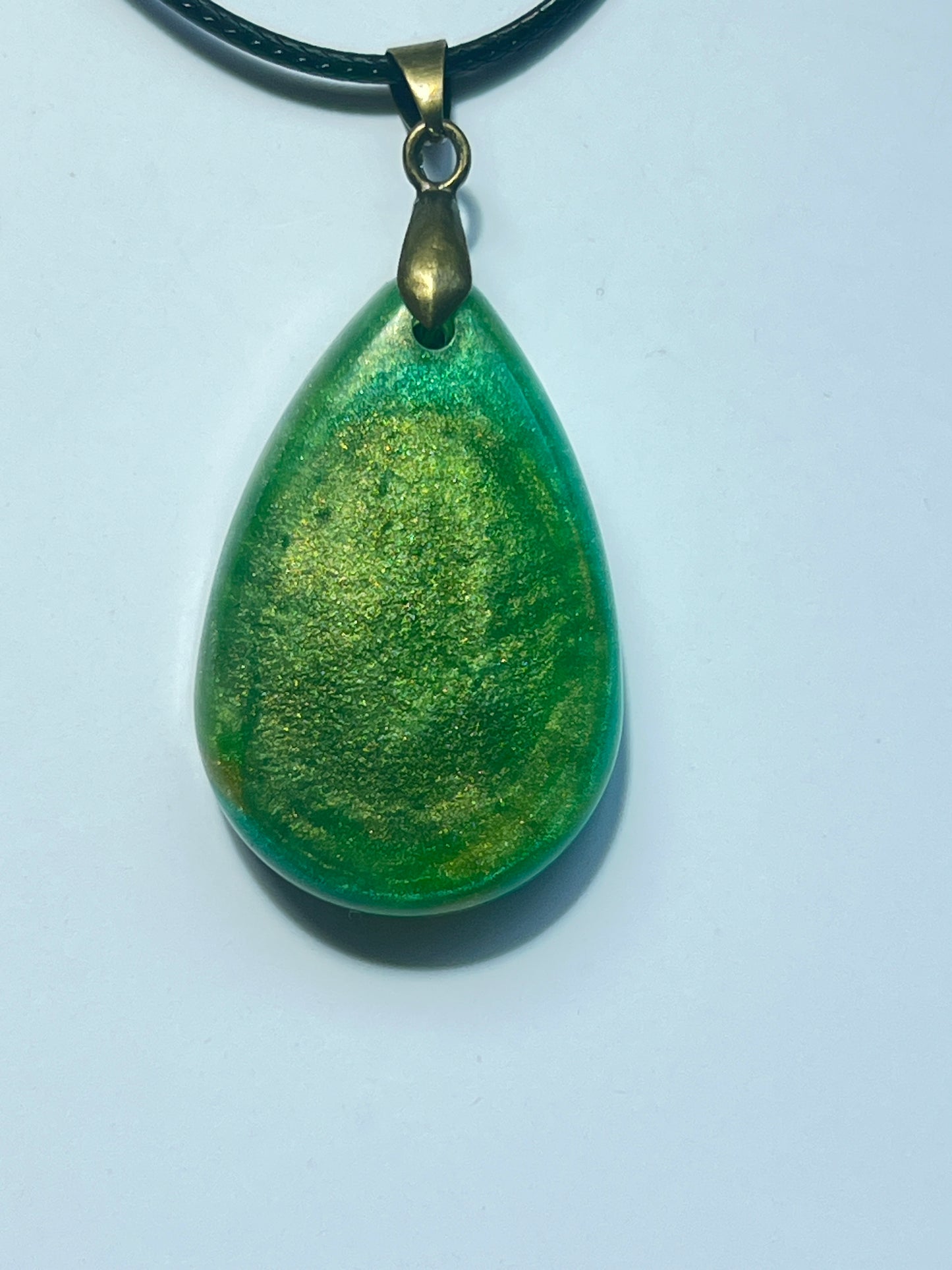 Handcrafted translucent emerald green resin wide teardrop pendant with swirling warm gold shimmer throughout giving a luminous inner glow effect, suspended from a black braided cord with an antique bronze bail.