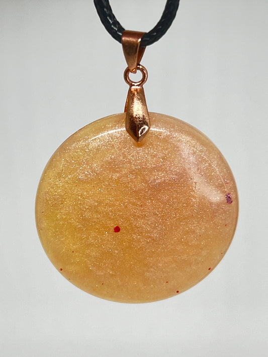 Handcrafted golden amber circle resin pendant with rose shimmer and scattered crimson flecks, suspended from a black braided cord with a copper-toned bail.