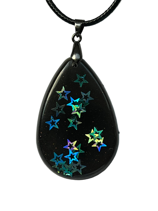 Handcrafted black resin teardrop pendant with holographic star confetti inclusions in teal, blue, purple, and gold, suspended from a black braided cord with a gunmetal bail.