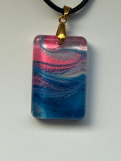Handcrafted rounded rectangle resin pendant with layered swirling hot pink, violet, white, and cobalt blue pigments in a wave pattern, suspended from a black braided cord with a gold-toned bail.