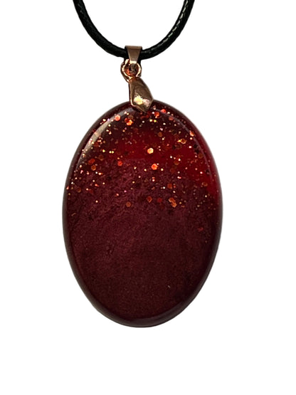 Handcrafted deep burgundy red resin oval pendant with scattered copper and orange glitter in the upper portion fading into a rich velvety matte base, suspended from a black braided cord with a copper-toned bail.