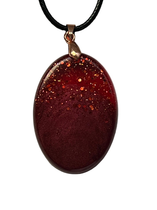 Handcrafted deep burgundy red resin oval pendant with scattered copper and orange glitter in the upper portion fading into a rich velvety matte base, suspended from a black braided cord with a copper-toned bail.