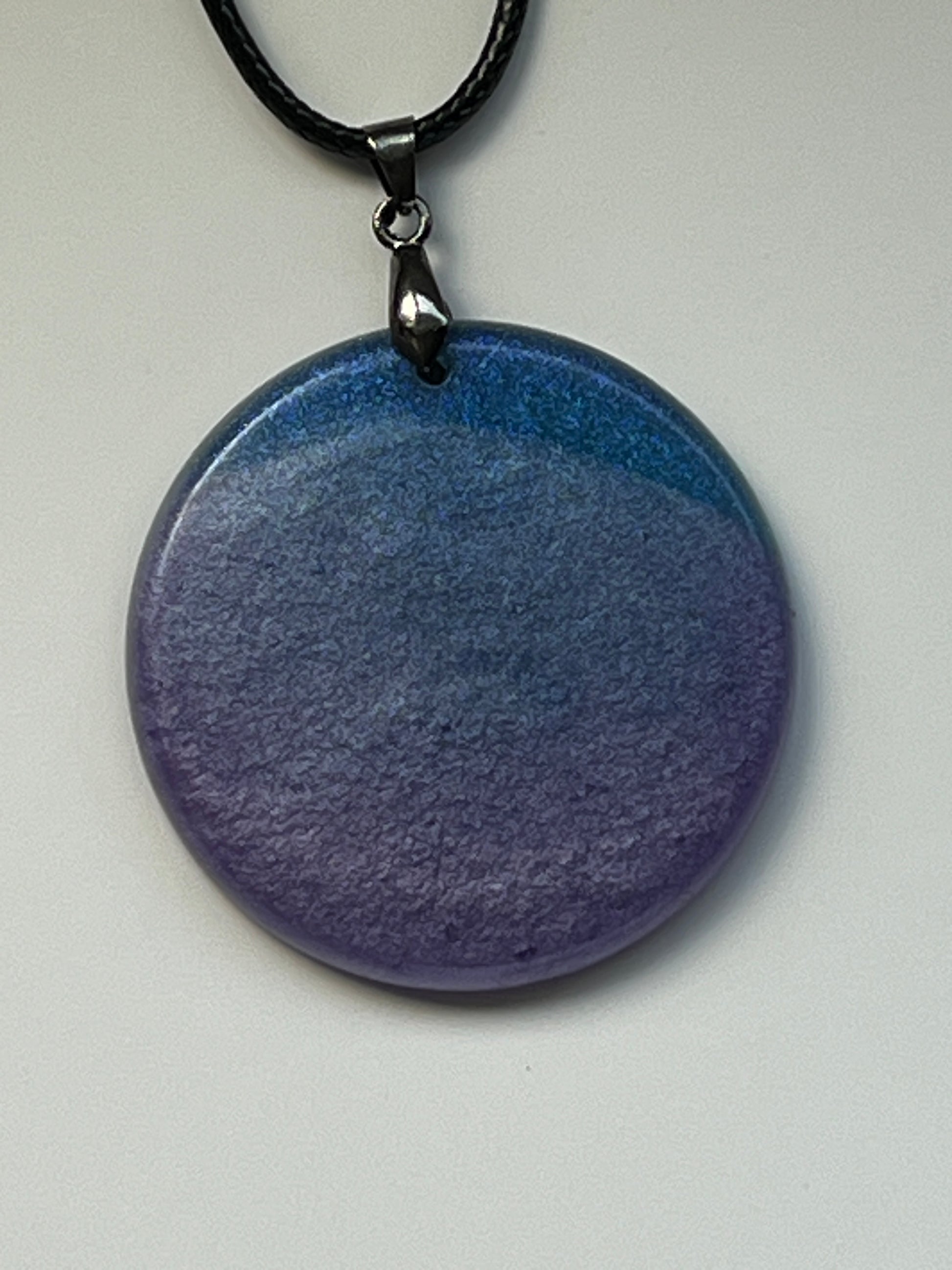Handcrafted resin large circle pendant with soft dusty blue upper tone fading into rich deep violet below with a fine matte mineral texture throughout, suspended from a black braided cord with a gunmetal bail.