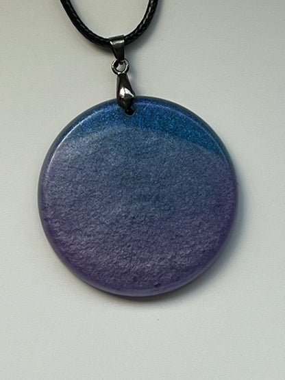 Handcrafted resin large circle pendant with soft dusty blue upper tone fading into rich deep violet below with a fine matte mineral texture throughout, suspended from a black braided cord with a gunmetal bail.