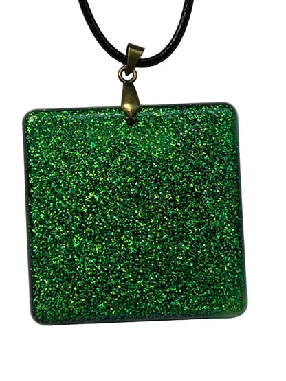 Handcrafted resin rounded square pendant packed with dense holographic emerald and forest green glitter with subtle darker swirl depth throughout, suspended from a black braided cord with an antique bronze bail.