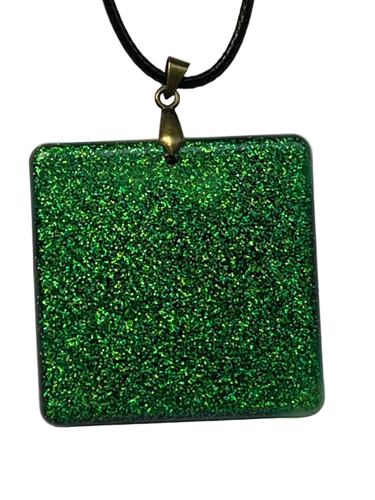 Handcrafted resin rounded square pendant packed with dense holographic emerald and forest green glitter with subtle darker swirl depth throughout, suspended from a black braided cord with an antique bronze bail.