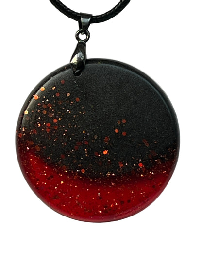 Handcrafted resin circle pendant with matte black upper half scattered with copper glitter transitioning into a blazing crimson red glitter base, suspended from a black braided cord with a gunmetal bail.