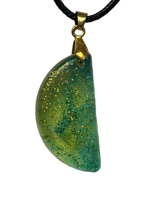 Handcrafted chartreuse green resin half moon pendant with dense holographic gold and teal glitter throughout, suspended from a black braided cord with a gold-toned bail.
