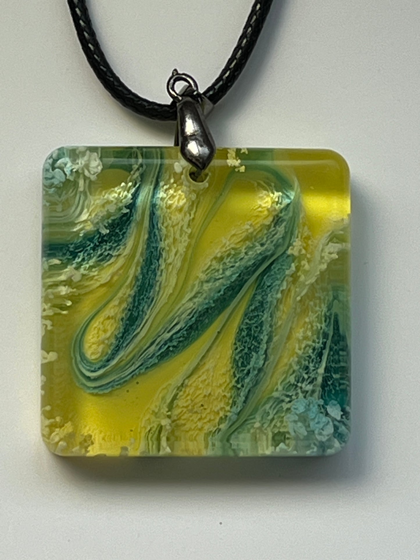 Handcrafted resin rounded square pendant with electric yellow base, sweeping deep teal and forest green swirls, and scattered white foam texture throughout, suspended from a black braided cord with a silver-toned bail.