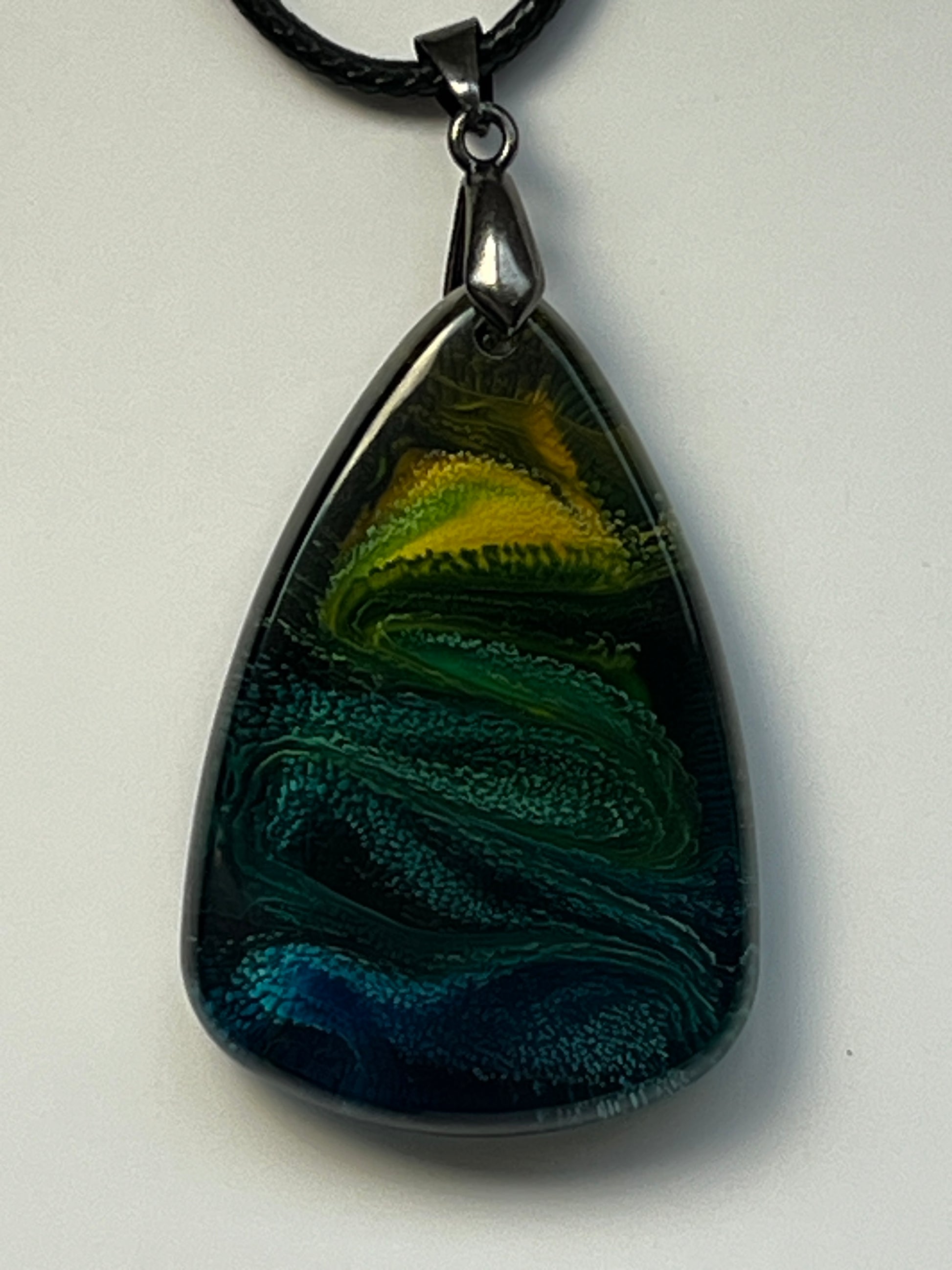 Handcrafted resin teardrop pendant with deep midnight navy and black base, sweeping teal and forest green wave texture, and a glowing electric yellow and chartreuse crown, suspended from a black braided cord with a gunmetal bail.