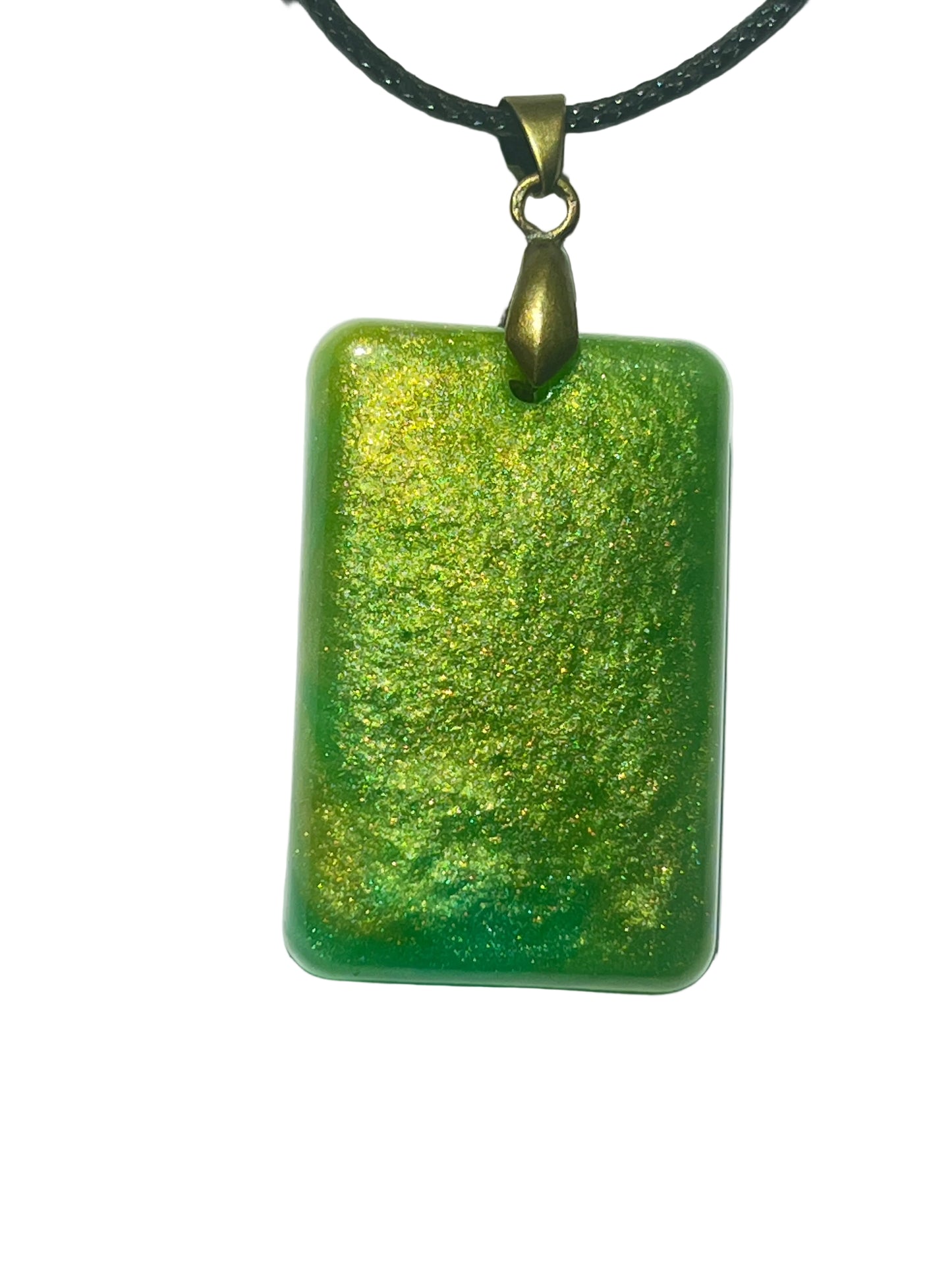 Handcrafted rich grass green resin rounded rectangle pendant with dense gold and chartreuse shimmer throughout creating a warm luminous meadow effect, suspended from a black braided cord with an antique bronze bail.