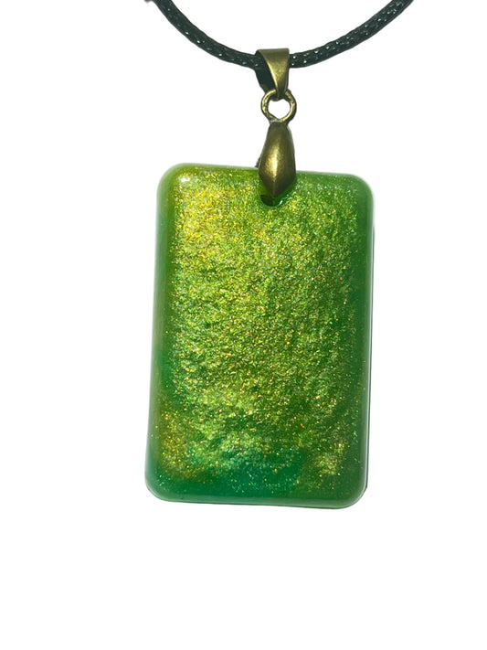 Handcrafted rich grass green resin rounded rectangle pendant with dense gold and chartreuse shimmer throughout creating a warm luminous meadow effect, suspended from a black braided cord with an antique bronze bail.