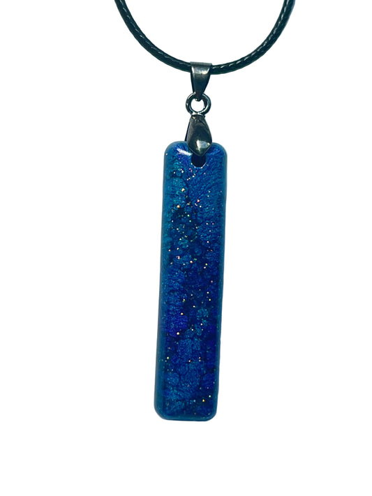 Handcrafted deep royal and cobalt blue resin elongated bar pendant with layered atmospheric texture and scattered holographic glitter in gold, copper, and teal throughout, suspended from a black braided cord with a silver-toned bail.