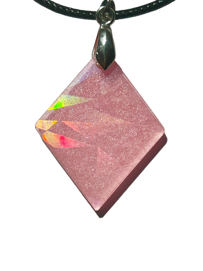 Handcrafted soft rose pink resin diamond-shaped pendant with fine silver glitter and holographic texture that fractures into rainbow light bursts in direct light, suspended from a black braided cord with a silver-toned bail.