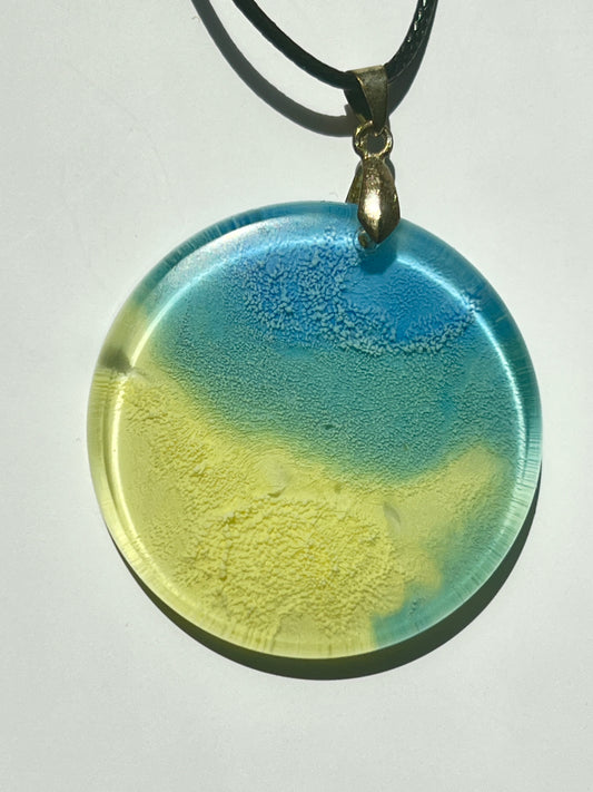Handcrafted resin large circle pendant with soft pale yellow-green lower field meeting periwinkle and sky blue upper tones with organic seafoam texture at the boundary, suspended from a black braided cord with an antique bronze bail.