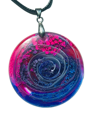 Handcrafted resin circle pendant with a three-dimensional relief spiral set against hot magenta pink with chunky glitter and deep cobalt blue swirls, suspended from a black braided cord with a silver-toned bail.