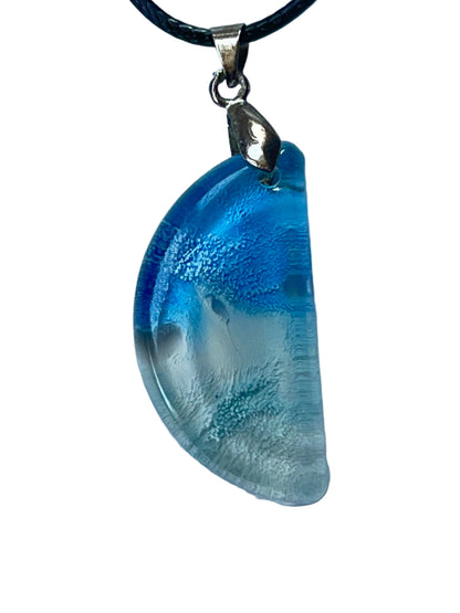 Handcrafted resin half moon pendant with vivid electric blue upper arc fading to pale aqua and near-total crystal clarity at the lower curved edge with fine horizontal texture, suspended from a black braided cord with a silver-toned bail.
