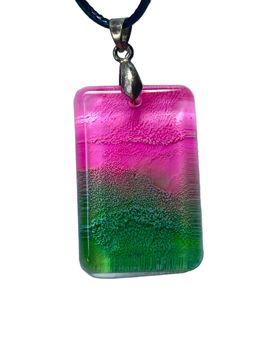 Handcrafted resin rounded rectangle pendant with hot magenta pink upper field, shimmering silver champagne middle band, and vivid grass green base with fine horizontal texture and blue glitter accents, suspended from a black braided cord with an antique bronze bail.