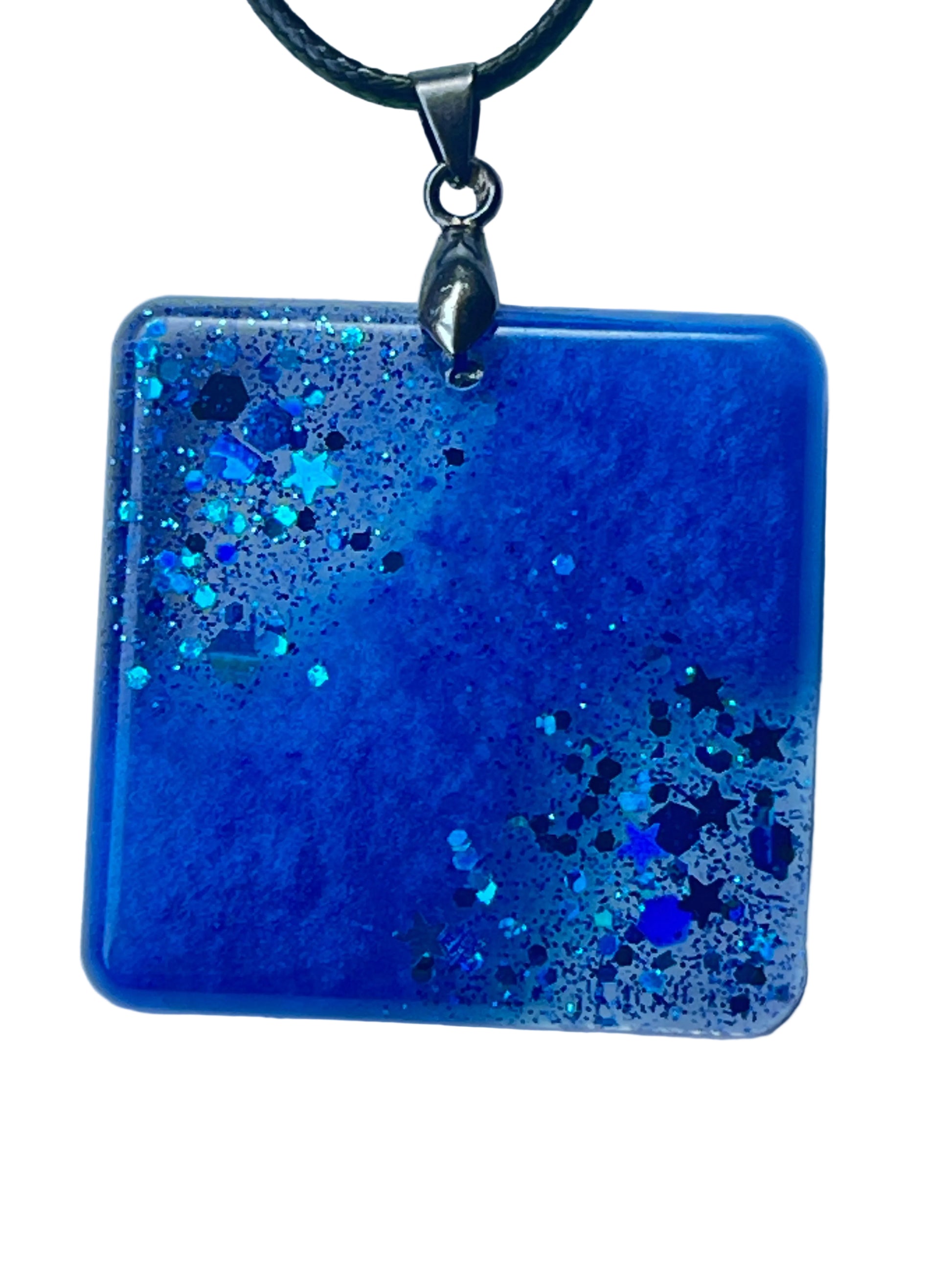 Handcrafted deep cobalt and royal blue resin rounded square pendant with a diagonal cascade of holographic star confetti, hexagon glitter, and fine teal and aqua shimmer across clear glassy resin, suspended from a black braided cord with a silver-toned bail.