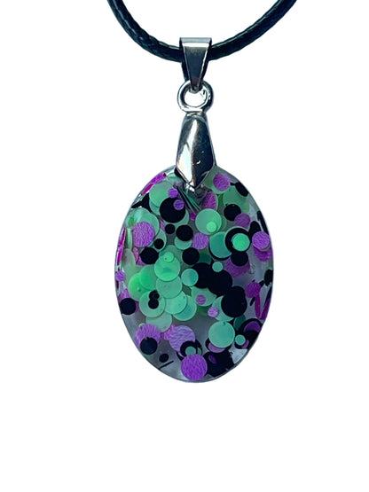 Handcrafted crystal clear resin oval pendant packed with chunky mint green, violet, and black circular confetti at multiple depths creating a three-dimensional cauldron bubble effect, suspended from a black braided cord with a silver-toned bail.