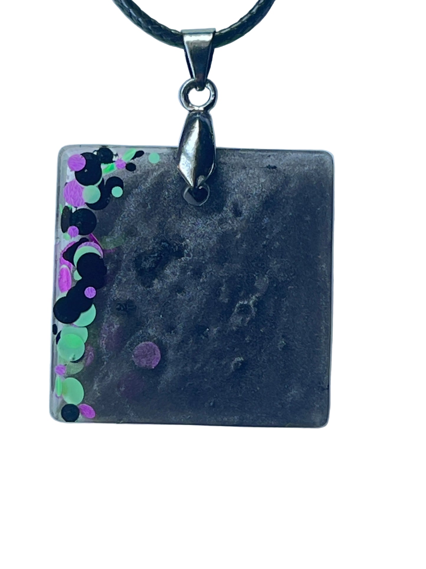 Handcrafted deep smoky charcoal and black resin rounded square pendant with a cascade of chunky mint green, violet, and black circular confetti spilling from the left edge into the dark mottled interior, suspended from a black braided cord with a silver-toned bail.
