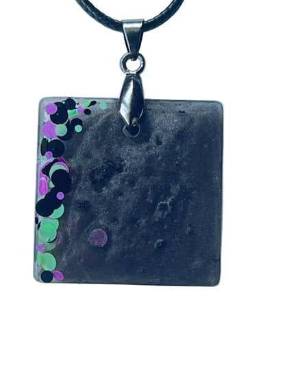 Handcrafted deep smoky charcoal and black resin rounded square pendant with a cascade of chunky mint green, violet, and black circular confetti spilling from the left edge into the dark mottled interior, suspended from a black braided cord with a silver-toned bail.