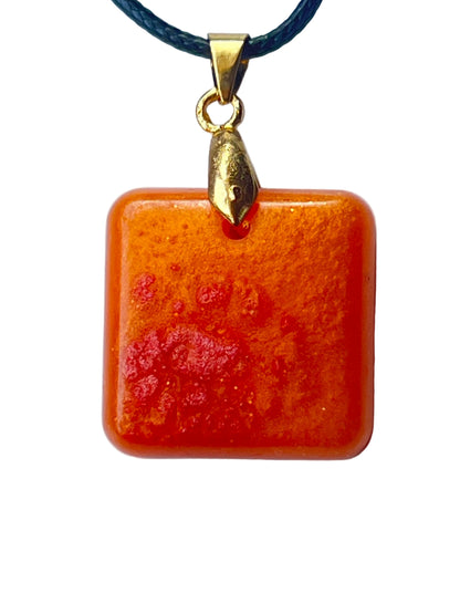 Handcrafted rich pumpkin and burnt orange resin rounded square pendant with fine gold shimmer and a deeper crimson red accent cluster, with a translucent jewel-like finish, suspended from a dark braided cord with a gold-toned bail.