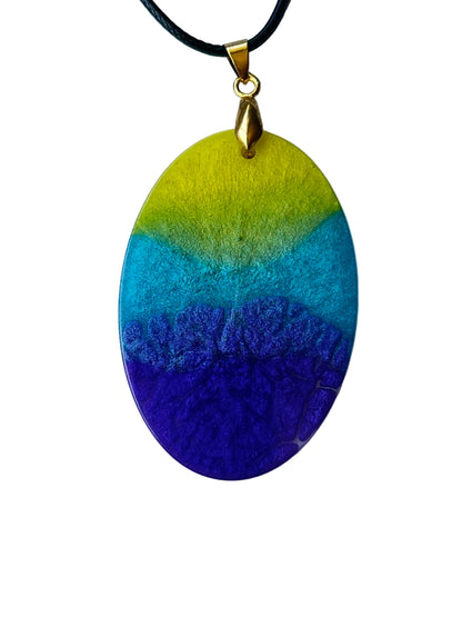 Handcrafted large resin oval pendant with bold solar chartreuse-yellow upper band, vivid aqua-teal center stripe, and rich deep blue-violet indigo base with fine complex texture throughout, suspended from a black braided cord with a gold-toned bail.
