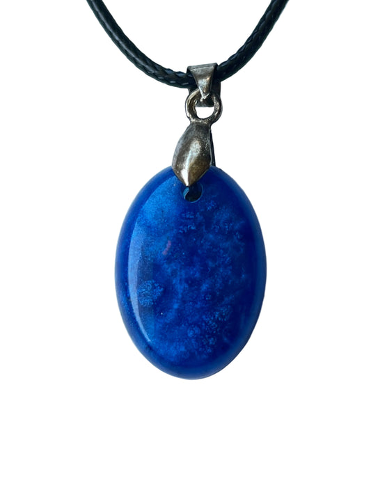 Handcrafted deep opaque royal blue resin oval pendant with natural organic tonal variation across the surface in the manner of polished lapis lazuli, with a high-gloss domed finish, suspended from a black braided cord with a silver-toned bail.