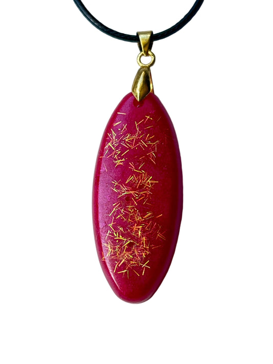 Handcrafted deep magenta and cranberry red resin elongated oval pendant with a central cascade of fine gold needle glitter catching warm metallic flashes, suspended from a black braided cord with a gold-toned bail.