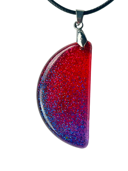 Handcrafted resin half-moon pendant with vivid translucent crimson upper field fading through magenta into deep violet and indigo lower arc, densely filled with red and electric blue fine glitter throughout, suspended from a black braided cord with a silver-toned bail.