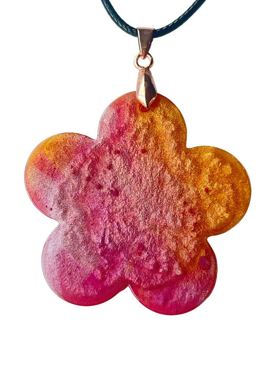 Handcrafted large resin five-petal flower pendant with vivid saturated magenta and hot pink center fading through peachy-coral into warm translucent amber-gold outer petal edges with fine botanical surface texture throughout, suspended from a black braided cord with a rose gold bail.