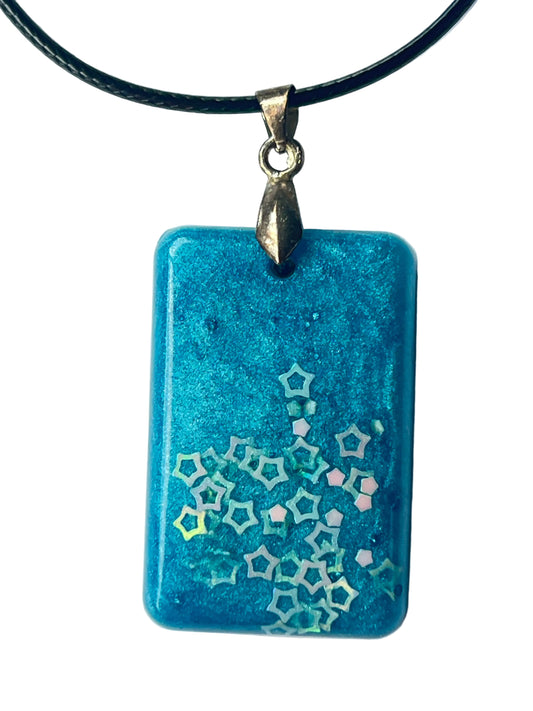 Handcrafted rich opaque teal resin rounded rectangle pendant with fine metallic shimmer throughout and an organic drifting cluster of iridescent hollow five-pointed star glitter pieces concentrated at the lower right and trailing upward across the surface, suspended from a black braided cord with an antiqued brass bail.