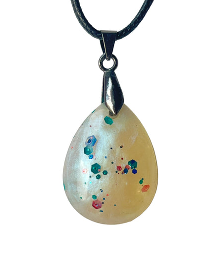 Handcrafted soft creamy ivory and pale yellow opalescent resin teardrop pendant with scattered multicolor glitter constellation including teal and emerald hexagonal flakes, coral and pink pieces, and electric blue fine glitter suspended at varying depths, suspended from a black braided cord with a gunmetal bail.