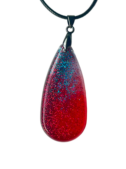 Handcrafted deep translucent crimson resin tall teardrop pendant with dense fine magenta-red glitter throughout and a dramatic bloom of electric teal and blue glitter in the upper field, suspended from a black braided cord with a gunmetal bail.