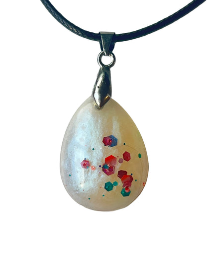 Handcrafted soft milky white opalescent resin teardrop pendant with pearlescent shimmer and a floating cluster of bold coral-orange and teal-emerald hexagonal glitter pieces suspended at varying depths, suspended from a black braided cord with a silver-toned bail.