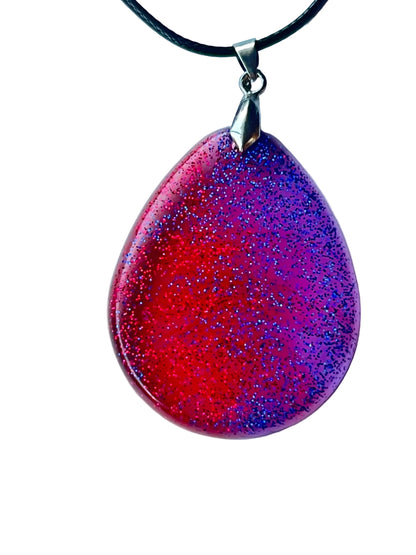 Handcrafted large resin teardrop pendant with vivid translucent crimson-red and electric violet fields meeting in a dynamic diagonal magenta transition, densely filled with ultra-fine red and blue-violet glitter throughout, suspended from a black braided cord with a silver-toned bail.