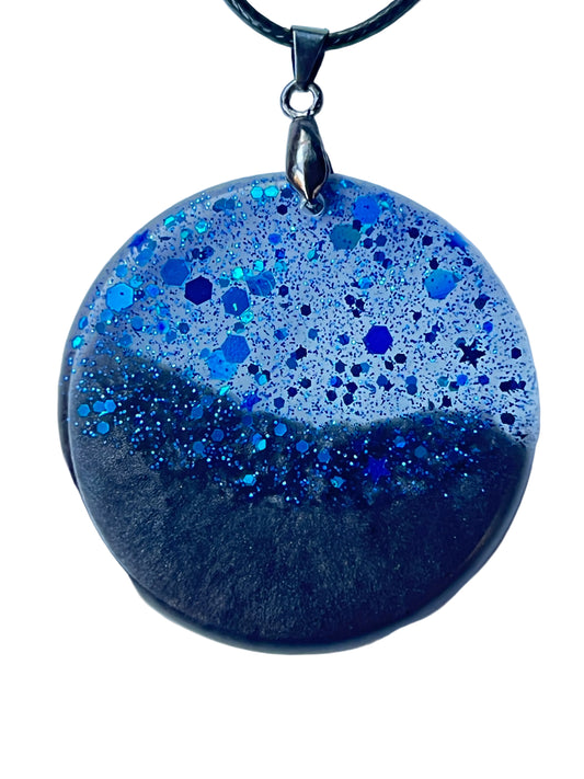 Alt text: Handcrafted large resin circle pendant with deep opaque black lower half and translucent silver-grey upper field densely packed with fine iridescent blue glitter and bold sapphire-blue hexagonal flakes meeting at a sweeping organic horizon line, suspended from a black braided cord with a gunmetal bail.