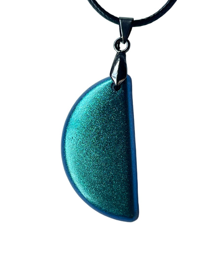 Handcrafted deep saturated teal resin half-moon pendant with extraordinarily fine dichroic metallic shimmer shifting between emerald-green and electric sapphire-blue throughout and a dark cobalt-blue flat edge, suspended from a black braided cord with a gunmetal bail.