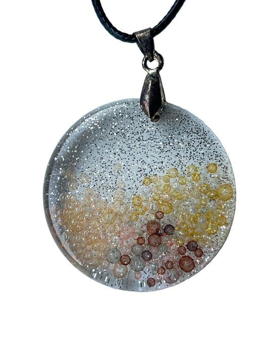 Handcrafted large resin circle pendant with clear crystal base, dense ultra-fine silver glitter haze in the upper field, and a micro-bubble landscape transitioning from golden-yellow through peachy amber to rust and copper-brown in the lower field, suspended from a black braided cord with a silver-toned bail.