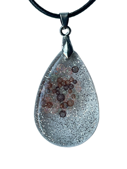 Handcrafted clear crystal resin teardrop pendant with extraordinarily dense ultra-fine silver glitter throughout and a floating cluster of micro-bubbles in blush-pink, peach, copper, and rust-brown suspended at varying depths in the upper-center field, suspended from a black braided cord with a silver-toned bail.