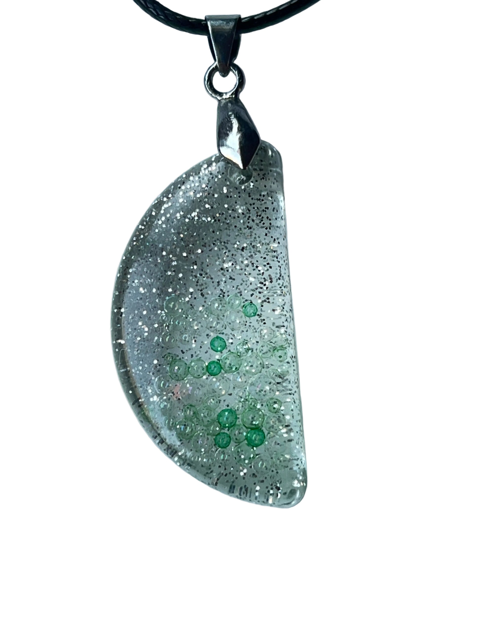 Handcrafted clear crystal resin half-moon pendant with extraordinarily dense ultra-fine silver glitter throughout and two organic clusters of bright emerald-green micro-bubbles floating at varying depths, suspended from a black braided cord with a silver-toned bail.

Next one whenever you're ready! 🌙✨