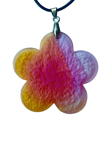 Handcrafted large resin five-petal flower pendant with warm translucent amber-gold outer edges flowing through peachy coral into deep saturated magenta-fuchsia center with fine surface texture throughout, suspended from a black braided cord with an antiqued gold bail.
