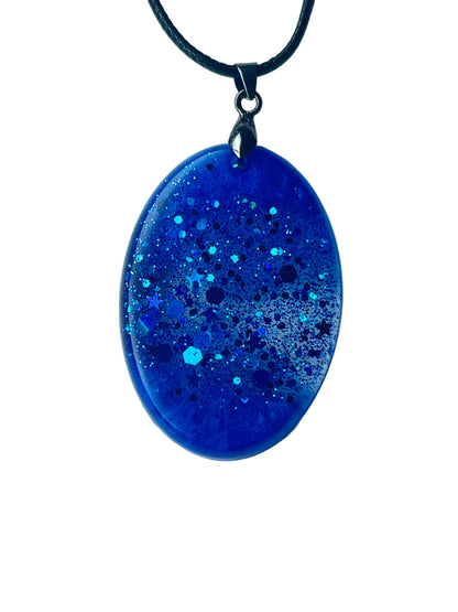 Handcrafted rich opaque royal blue resin oval pendant with a mixed glitter constellation of bold electric teal and sapphire hexagonal flakes, fine iridescent particles, and scattered five-pointed star shapes throughout, suspended from a black braided cord with a gunmetal bail.