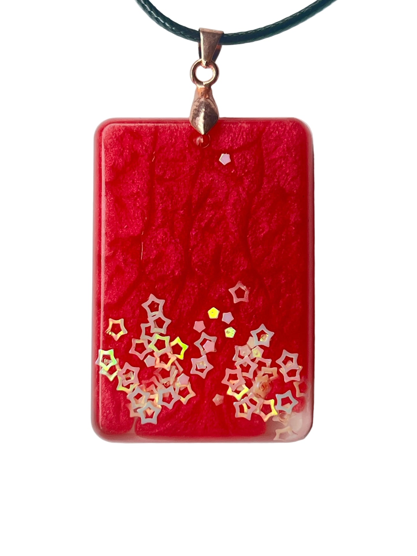 Handcrafted rich opaque crimson resin rounded rectangle pendant with intricate organic surface texture and a rising constellation of iridescent hollow five-pointed star glitter pieces densest at the lower corners and trailing upward, suspended from a black braided cord with a rose gold bail.