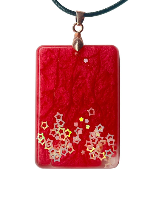 Handcrafted rich opaque crimson resin rounded rectangle pendant with intricate organic surface texture and a rising constellation of iridescent hollow five-pointed star glitter pieces densest at the lower corners and trailing upward, suspended from a black braided cord with a rose gold bail.