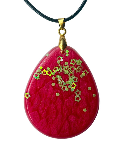 Handcrafted deep saturated magenta-crimson resin large teardrop pendant with intricate organic surface texture and a swirling cluster of iridescent gold and chartreuse hollow five-pointed star glitter pieces densest at upper center and scattering outward, suspended from a black braided cord with a gold-toned bail.
