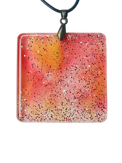 Handcrafted large resin square pendant in soft translucent coral-pink and warm peach with a golden-amber upper center and dense mixed red, crimson, and silver iridescent glitter throughout, suspended from a black braided cord with an antiqued brass bail.