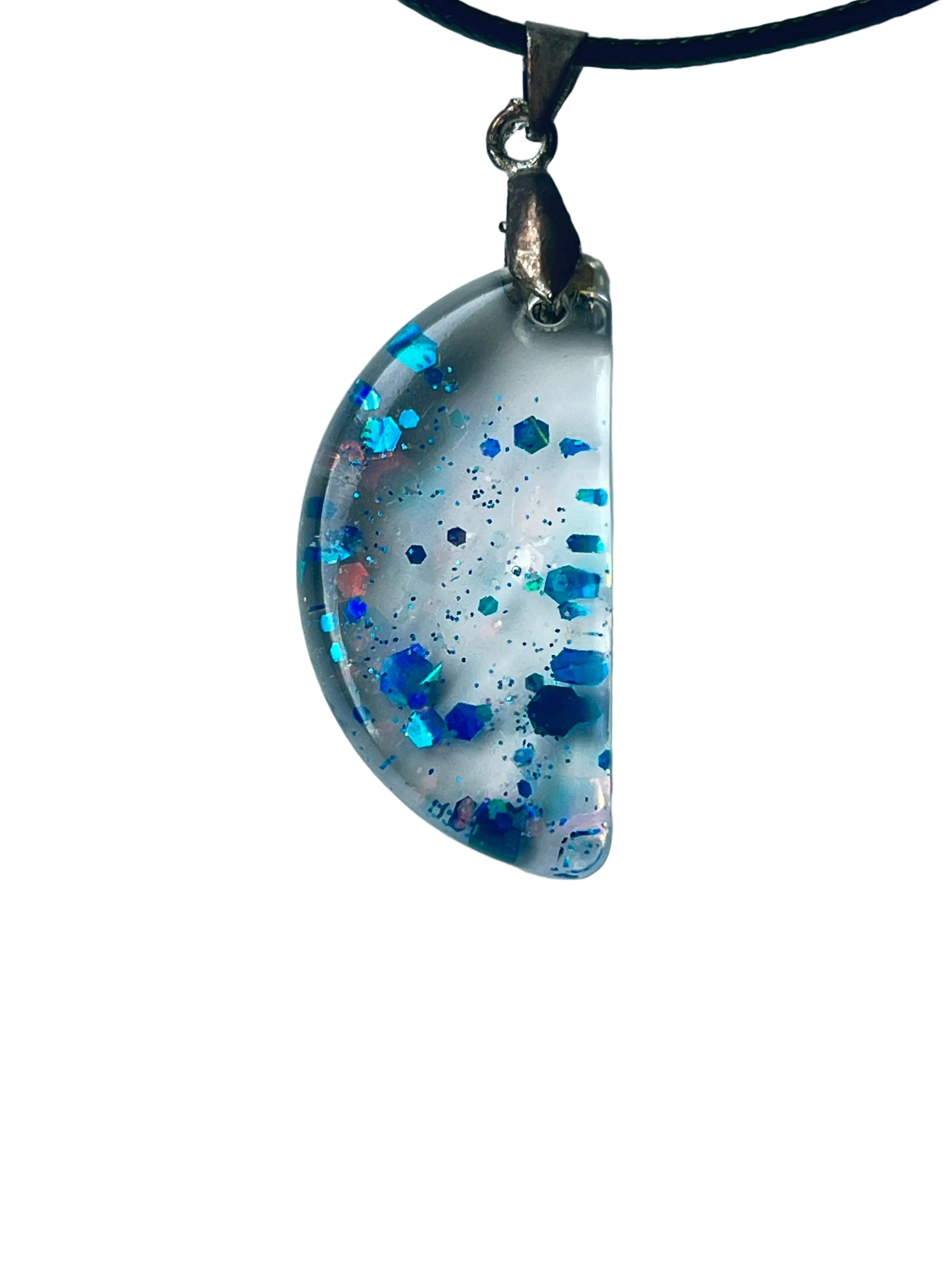 Handcrafted clear crystal resin half-moon pendant with scattered bold hexagonal glitter in electric blue, sapphire, and iridescent teal at varying depths with fine dark blue particle mist throughout, suspended from a black braided cord with a silver-toned bail.