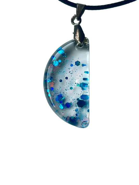 Handcrafted clear crystal resin half-moon pendant with scattered bold hexagonal glitter in electric blue, sapphire, and iridescent teal at varying depths with fine dark blue particle mist throughout, suspended from a black braided cord with a silver-toned bail.